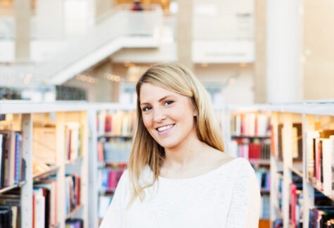 Smiling woman holding books in a sunny library setting.