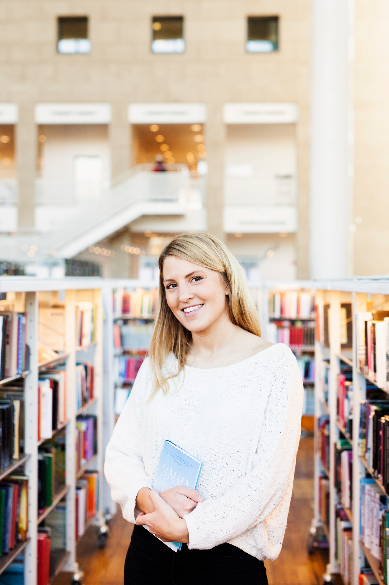Smiling woman holding books in a sunny library setting.